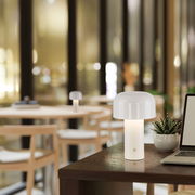 Moderne Akku-Tischleuchte AGNES in einem stilvollen Innenraum, auf einem Tisch neben einem Laptop und einer Pflanze platziert, mit Blick auf einen gemütlichen Raum im Hintergrund.