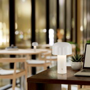 Moderne Akku-Tischleuchte AGNES in einem stilvollen Innenraum, auf einem Tisch neben einem Laptop und einer Pflanze platziert, mit Blick auf einen gemütlichen Raum im Hintergrund.