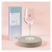 Weinglas mit Smiley-Design, frontal auf einem runden Sockel vor einer Verpackung präsentiert.