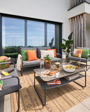 Moderne Loungegarnitur auf einer Terrasse mit grauen Sofas, bunten Kissen und einem Tisch mit Dekorationen, aus der Perspektive von vorne.