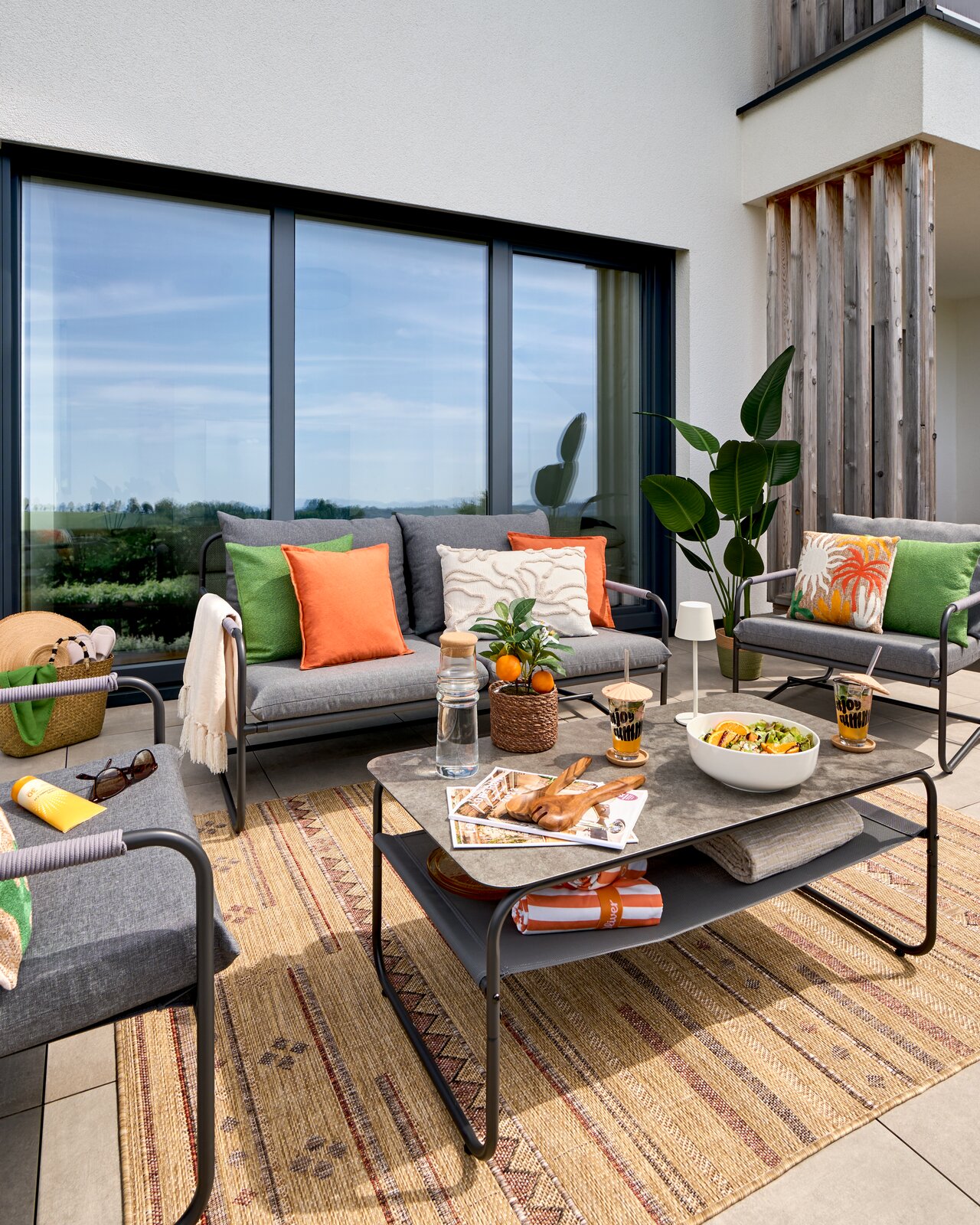 Moderne Loungegarnitur auf einer Terrasse mit grauen Sofas, bunten Kissen und einem Tisch mit Dekorationen, aus der Perspektive von vorne.