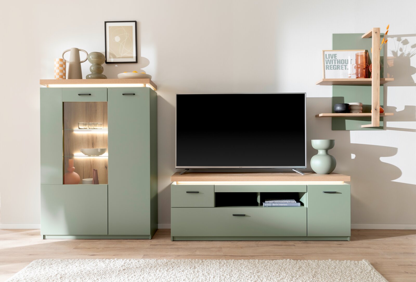 Frontale Ansicht einer modernen Wohnzimmermöbel-Kombination in Salbeigrün mit Holzoptik: Vitrine mit beleuchteten Glasböden, TV-Lowboard mit großem Fernseher und wandmontiertem Regal.