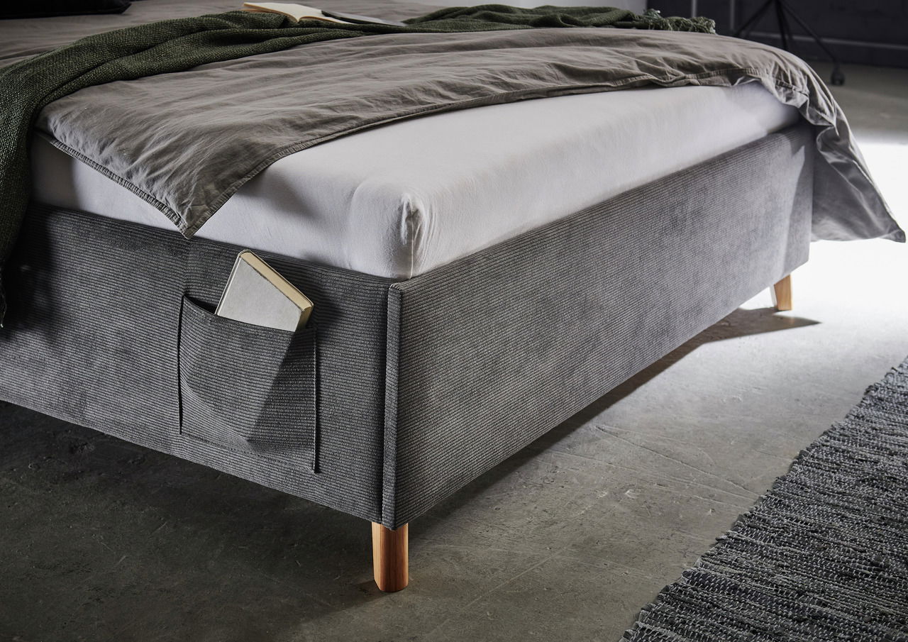 Seitliche Perspektive einer grauen Polsterliege mit Holzfüßen und einer Tasche an der Seite, die ein Buch enthält.