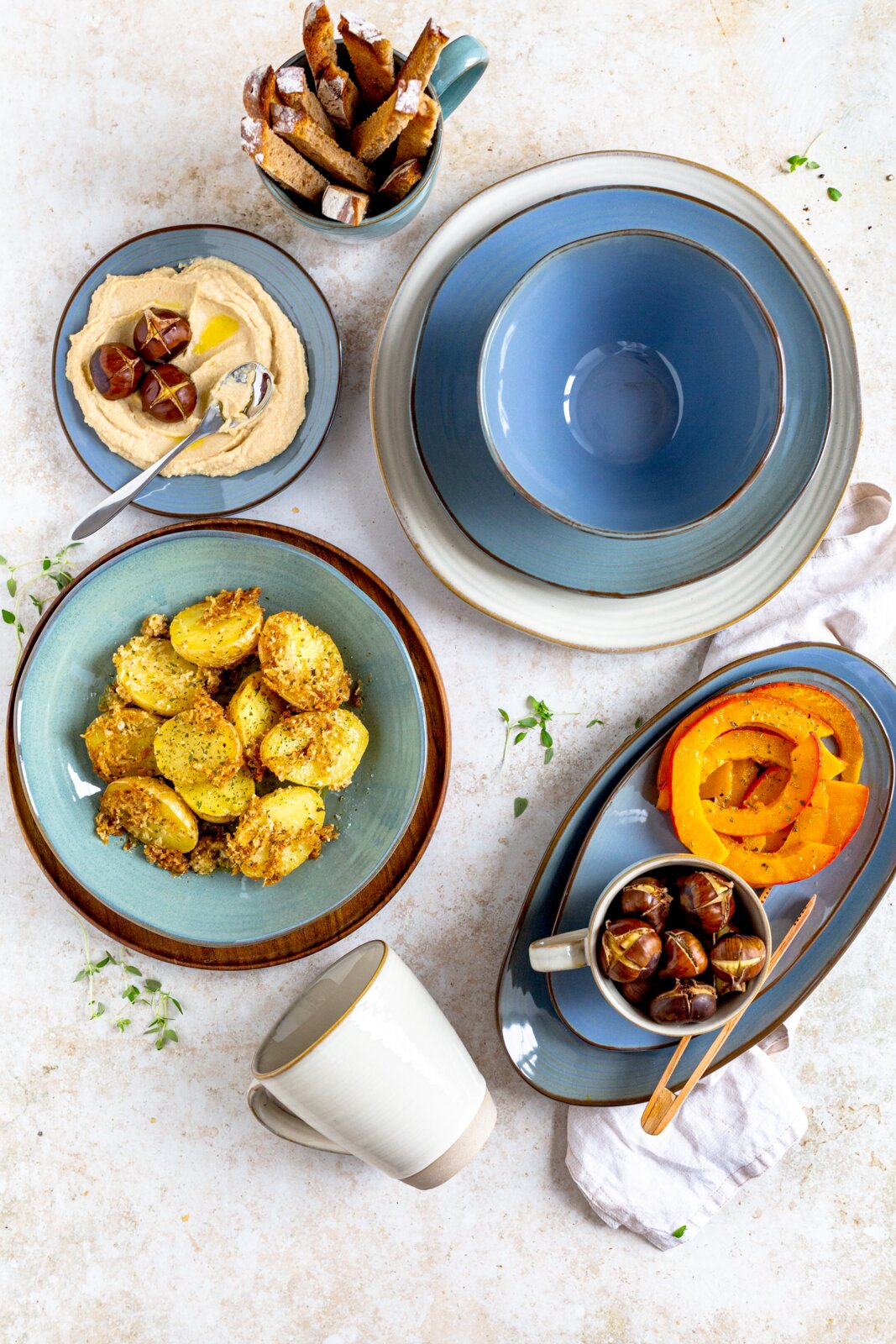 Draufsicht auf ein blau-graues Keramikgeschirr-Set mit tiefem Teller und ovaler Platte, arrangiert mit gerösteten Kartoffeln, Kürbisscheiben, Hummus, Esskastanien und Brotsticks auf hellem Tisch