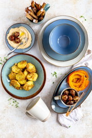 Draufsicht auf ein blau-graues Keramikgeschirr-Set mit tiefem Teller und ovaler Platte, arrangiert mit gerösteten Kartoffeln, Kürbisscheiben, Hummus, Esskastanien und Brotsticks auf hellem Tisch