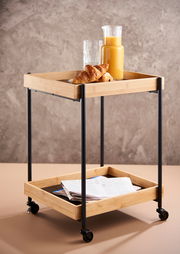 Moderner Servierwagen mit zwei Ablagen aus Holz und Metall, gefüllt mit einem Krug Orangensaft, Gläsern und Croissants, auf Rollen, seitliche Perspektive.