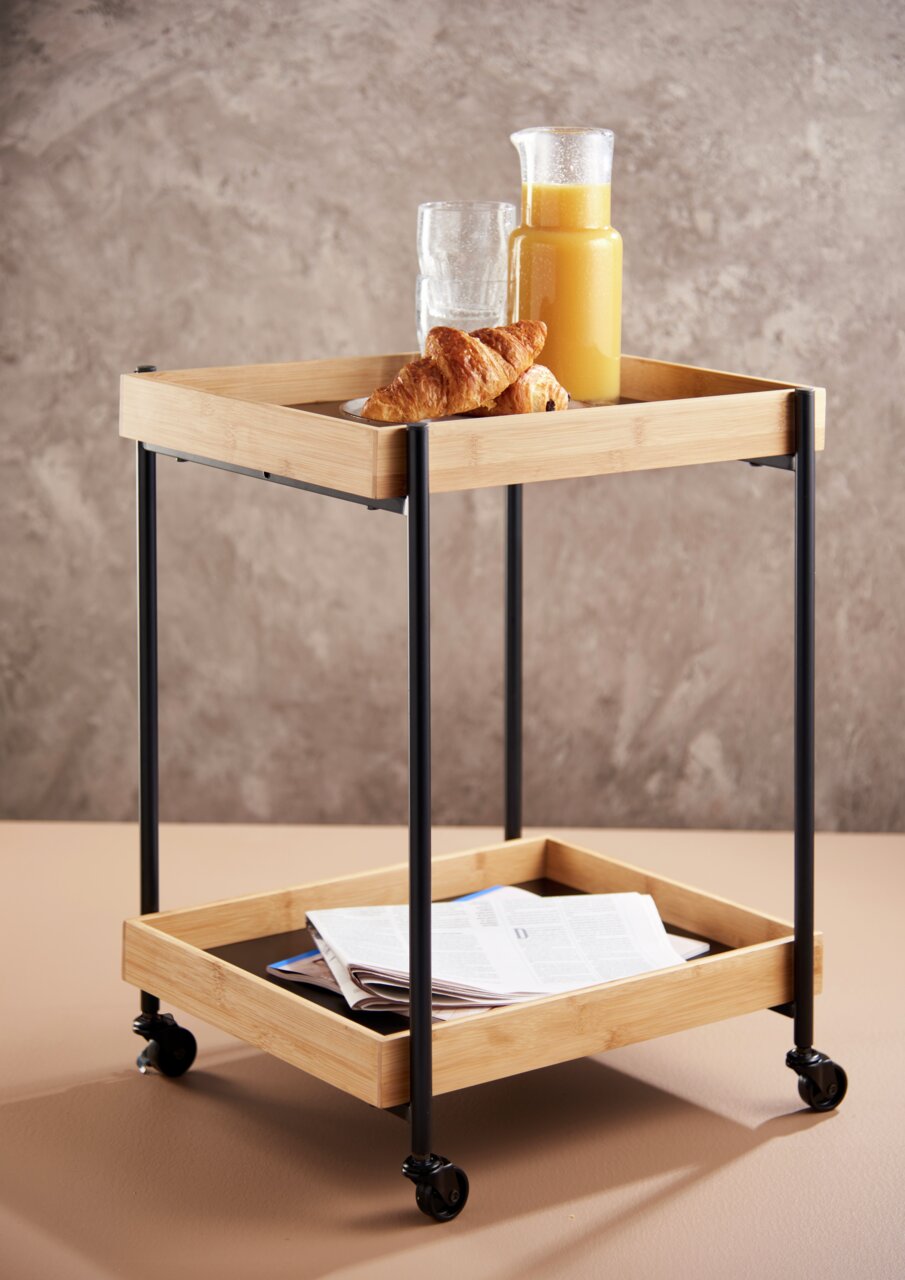 Moderner Servierwagen mit zwei Ablagen aus Holz und Metall, gefüllt mit einem Krug Orangensaft, Gläsern und Croissants, auf Rollen, seitliche Perspektive.