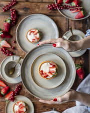 Draufsicht (Top-Down) auf eine Hand, die einen grauen Keramikteller mit Erdbeer-Tartelette und Vanillecreme über einem rustikalen Holztisch mit weiteren Tellern, Löffeln und frischen Beeren hält.