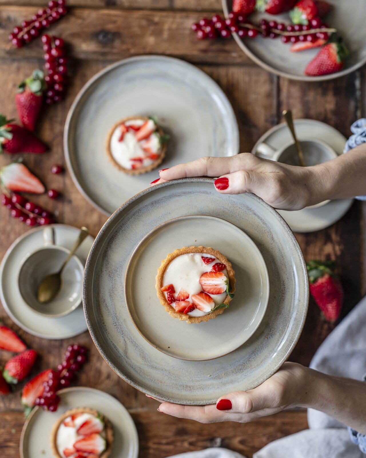 Draufsicht (Top-Down) auf eine Hand, die einen grauen Keramikteller mit Erdbeer-Tartelette und Vanillecreme über einem rustikalen Holztisch mit weiteren Tellern, Löffeln und frischen Beeren hält.