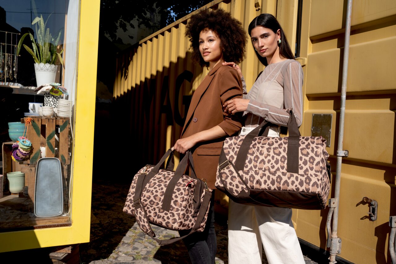 Reisenthel Allrounder L LEO MACCHIATO Zwei Frauen halten Leo Macchiato Allrounder Taschen vor einem gelben Container. Die Taschen haben ein Leopardenmuster und sind aus der Perspektive von vorne zu sehen.