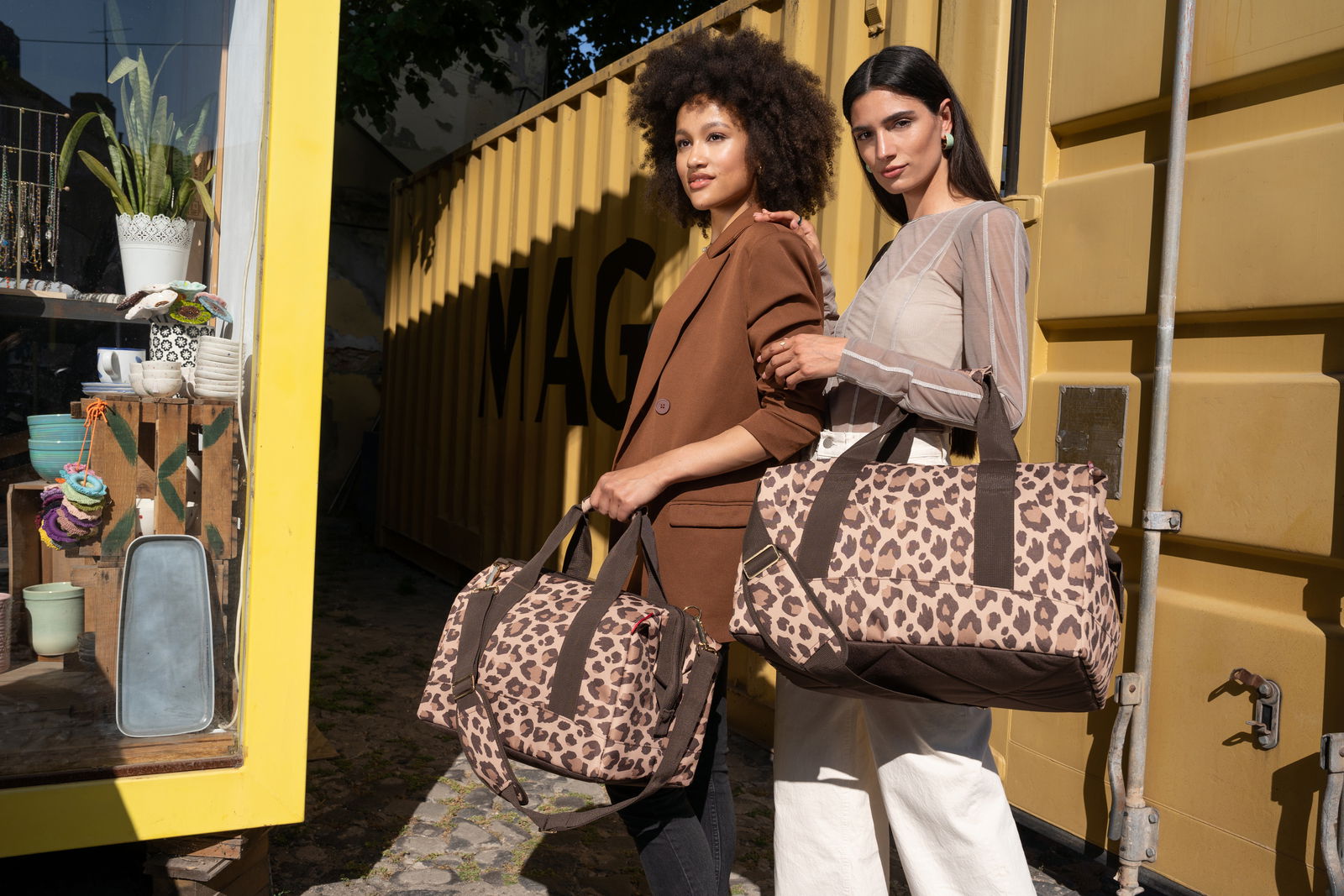 Zwei Frauen halten Leo Macchiato Allrounder Taschen vor einem gelben Container. Die Taschen haben ein Leopardenmuster und sind aus der Perspektive von vorne zu sehen.