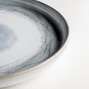 Nahaufnahme eines runden Keramiktellers in Blau-Grau mit gesprenkelter Glasur und dunklem Rand, schräg von oben fotografiert.
