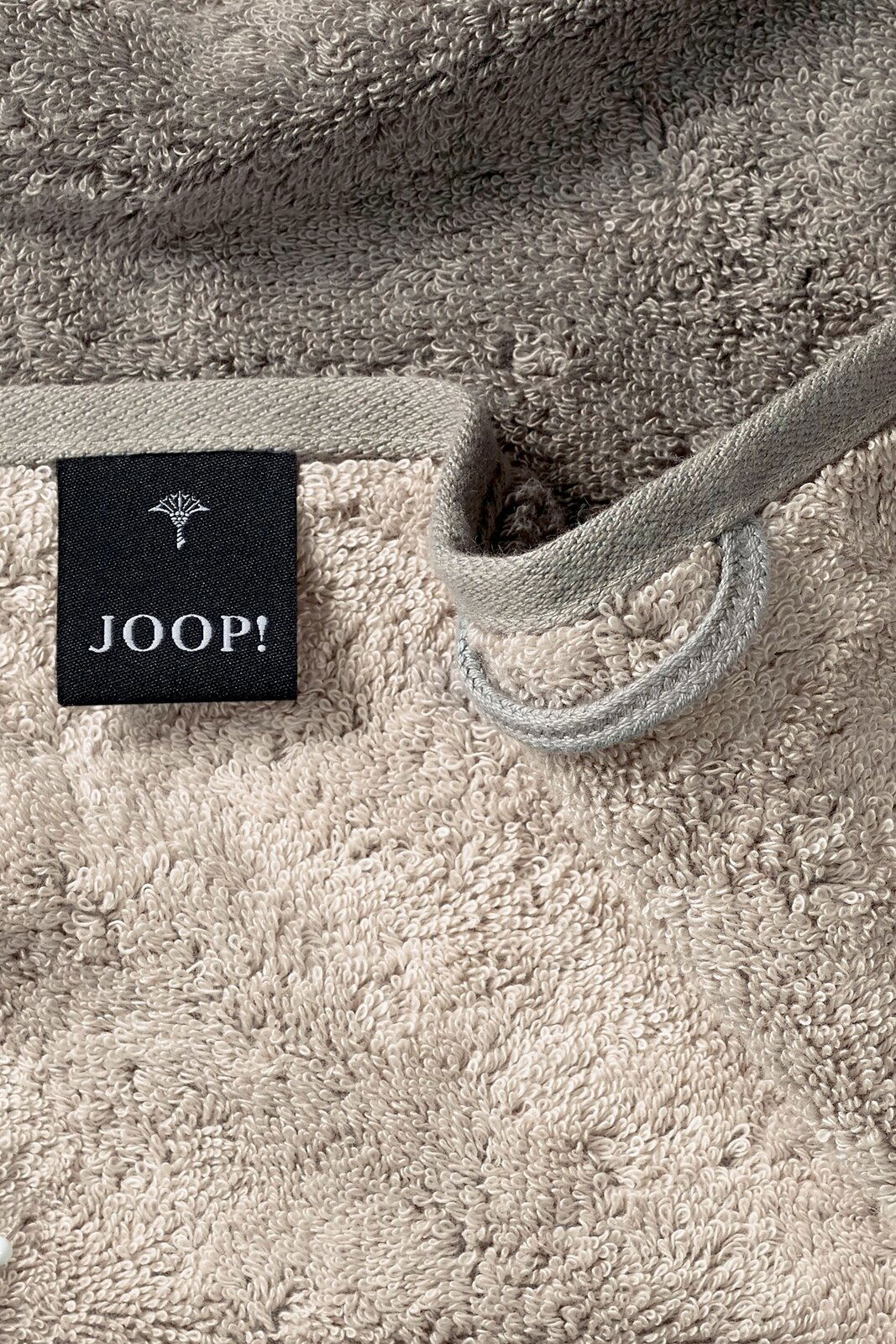 Nahaufnahme eines beigen Gästetuchs mit einer schwarzen Etikette mit der Aufschrift 'JOOP!' und einer Schlaufe zum Aufhängen.