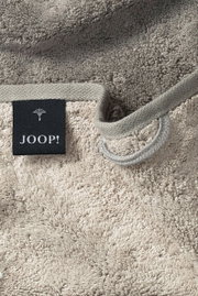 Nahaufnahme eines beigen Gästetuchs mit einer schwarzen Etikette mit der Aufschrift 'JOOP!' und einer Schlaufe zum Aufhängen.