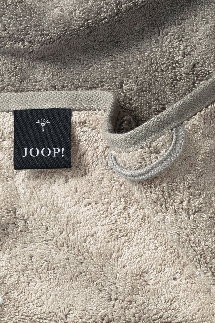 Nahaufnahme eines beigen Gästetuchs mit einer schwarzen Etikette mit der Aufschrift 'JOOP!' und einer Schlaufe zum Aufhängen.
