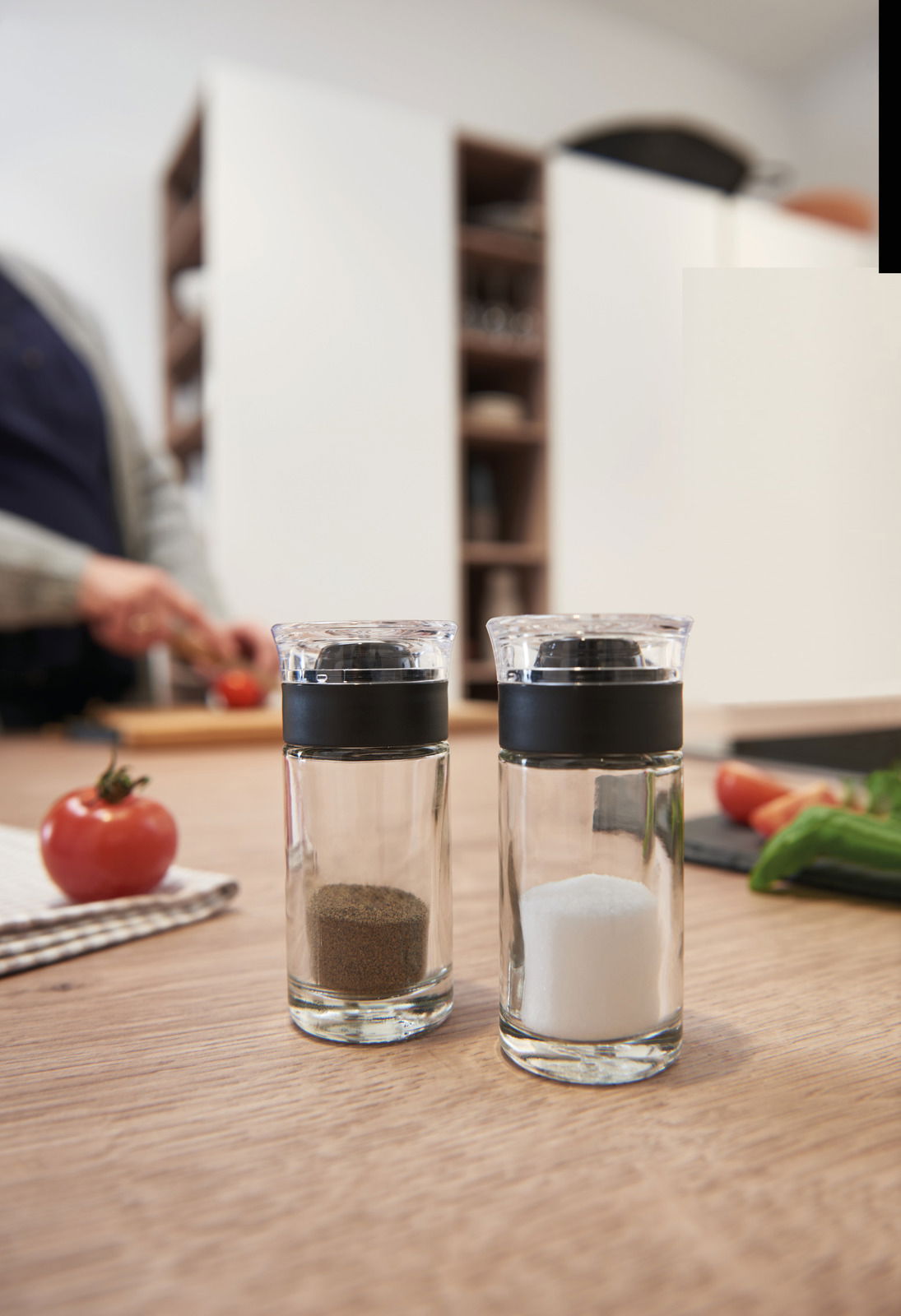 Leonardo Salz- & Pfefferstreuer 2-tlg. CUCINA Salz- und Pfefferstreuer Cucina aus Glas mit schwarzem Deckel, auf einem Holztisch in einer Küche, im Hintergrund eine Person beim Schneiden von Gemüse.
