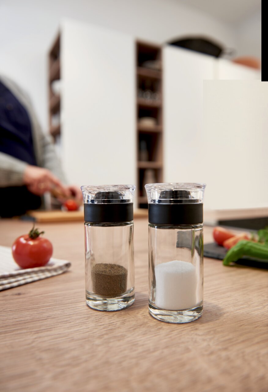 Salz- und Pfefferstreuer Cucina aus Glas mit schwarzem Deckel, auf einem Holztisch in einer Küche, im Hintergrund eine Person beim Schneiden von Gemüse.