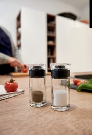 Salz- und Pfefferstreuer Cucina aus Glas mit schwarzem Deckel, auf einem Holztisch in einer Küche, im Hintergrund eine Person beim Schneiden von Gemüse.