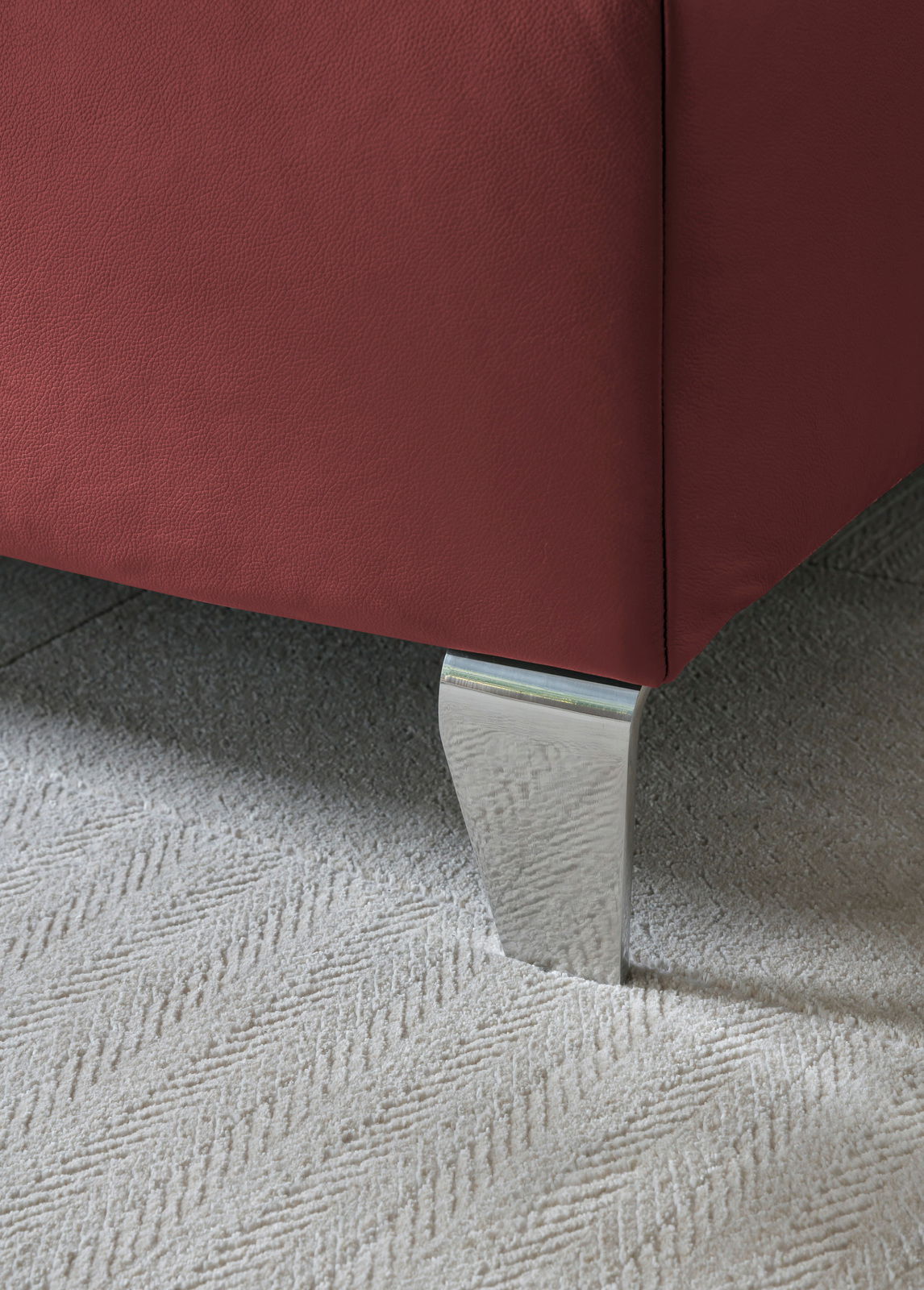 Nahaufnahme eines Boxspringbettes mit rotem Stoffbezug und silbernem Metallfuß auf Teppichboden, Perspektive von unten.