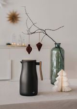 Schwarze Isolierkanne Studio Allure mit Holzgriff, seitliche Perspektive, auf einem Tisch mit dekorativen Elementen im Hintergrund.