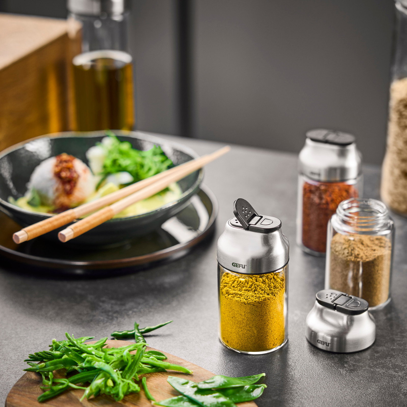 GEFU Gewürz- / Kräuterstreuer KIPPO Vorderansicht eines modernen Gewürz- und Kräuterstreuers aus Edelstahl und Glas, gefüllt mit gelbem Gewürz, umgeben von weiteren Gewürzgläsern und frischen Zutaten auf einem Küchentisch.