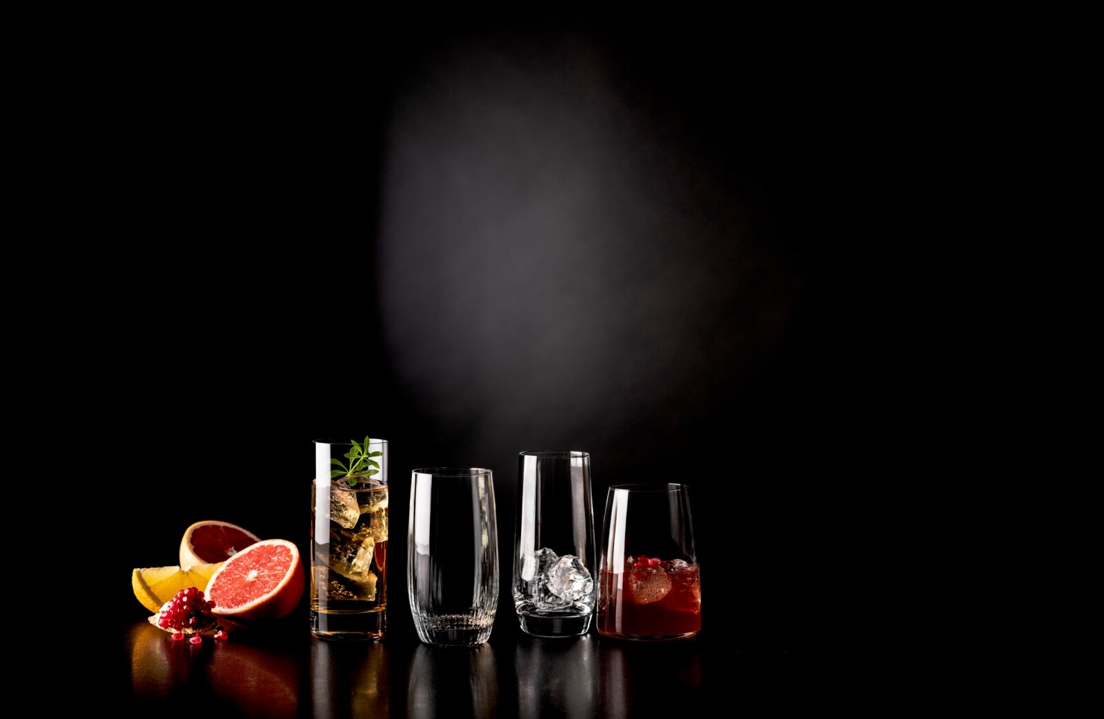 Stillleben aus vier transparenten Trinkgläsern mit Cocktails und Eiswürfeln neben Grapefruit, Zitronenscheibe und Granatapfelkernen auf schwarzem, spiegelndem Untergrund vor dunklem Hintergrund, fotografiert in frontaler Perspektive auf Augenhöhe.