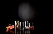 Stillleben aus vier transparenten Trinkgläsern mit Cocktails und Eiswürfeln neben Grapefruit, Zitronenscheibe und Granatapfelkernen auf schwarzem, spiegelndem Untergrund vor dunklem Hintergrund, fotografiert in frontaler Perspektive auf Augenhöhe.