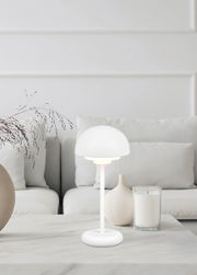 Moderne Akku-Tischleuchte ELLIOT in Weiß auf einem Tisch in einem stilvollen Wohnzimmer, seitliche Perspektive.