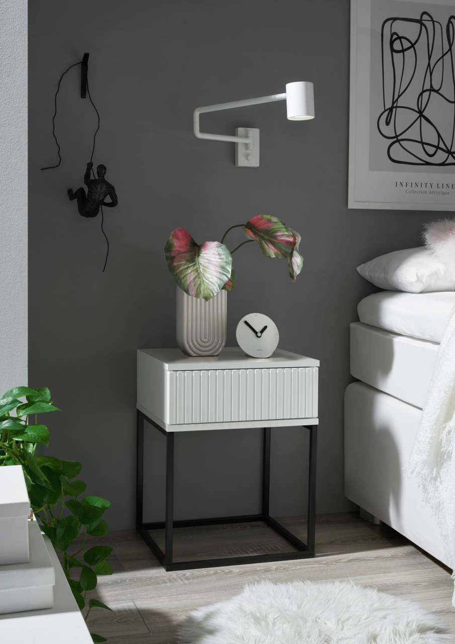 Moderne Nachtkommode mit weißer Schublade und schwarzem Metallgestell, seitliche Perspektive, dekoriert mit einer Pflanze und einer Uhr.