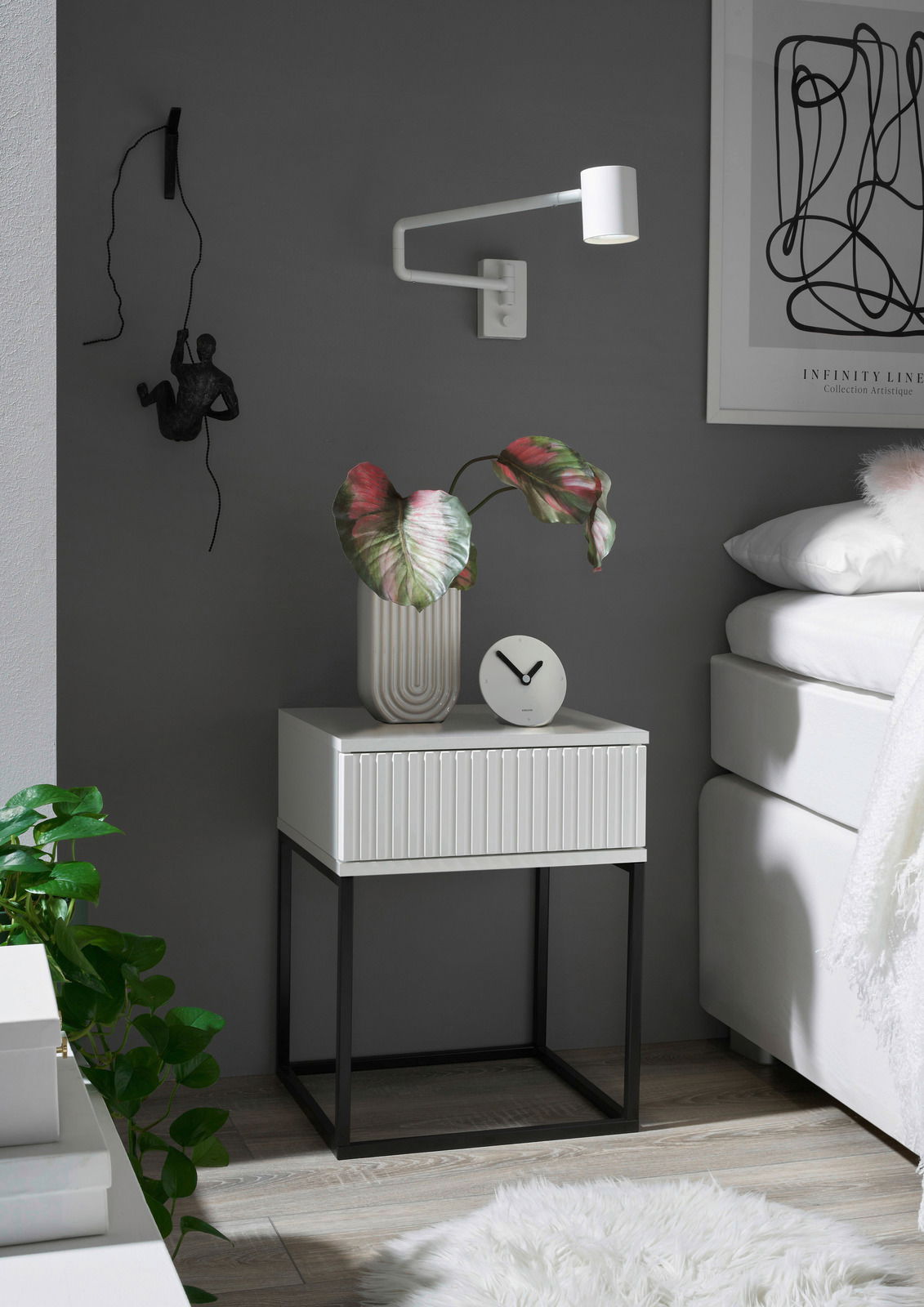 Moderne Nachtkommode mit weißer Schublade und schwarzem Metallgestell, seitliche Perspektive, dekoriert mit einer Pflanze und einer Uhr.