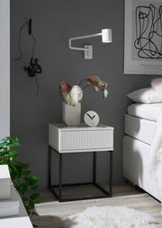 Moderne Nachtkommode mit weißer Schublade und schwarzem Metallgestell, seitliche Perspektive, dekoriert mit einer Pflanze und einer Uhr.