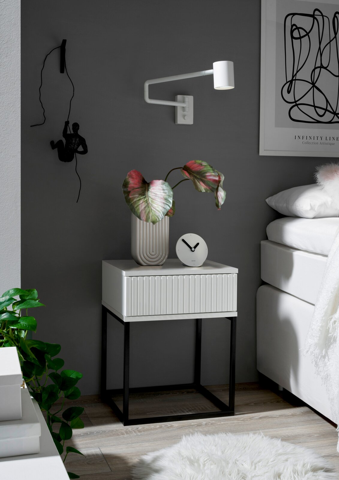 Moderne nachtkastje met witte lade en zwart metalen onderstel, gezien vanaf de zijkant, versierd met een plant en een klok.