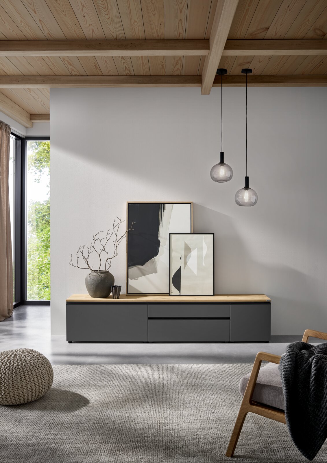 Modernes, niedriges Sideboard/TV-Lowboard in Anthrazit mit heller Holzplatte, frontale Ansicht in einem minimalistischen Wohnzimmer darauf zwei gerahmte Bilder und eine Vase, darüber zwei Glas-Pendelleuchten.