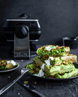 Waffeleisen Professional in der Seitenansicht mit einem Teller voller grüner Waffeln im Vordergrund, garniert mit weißer Soße.