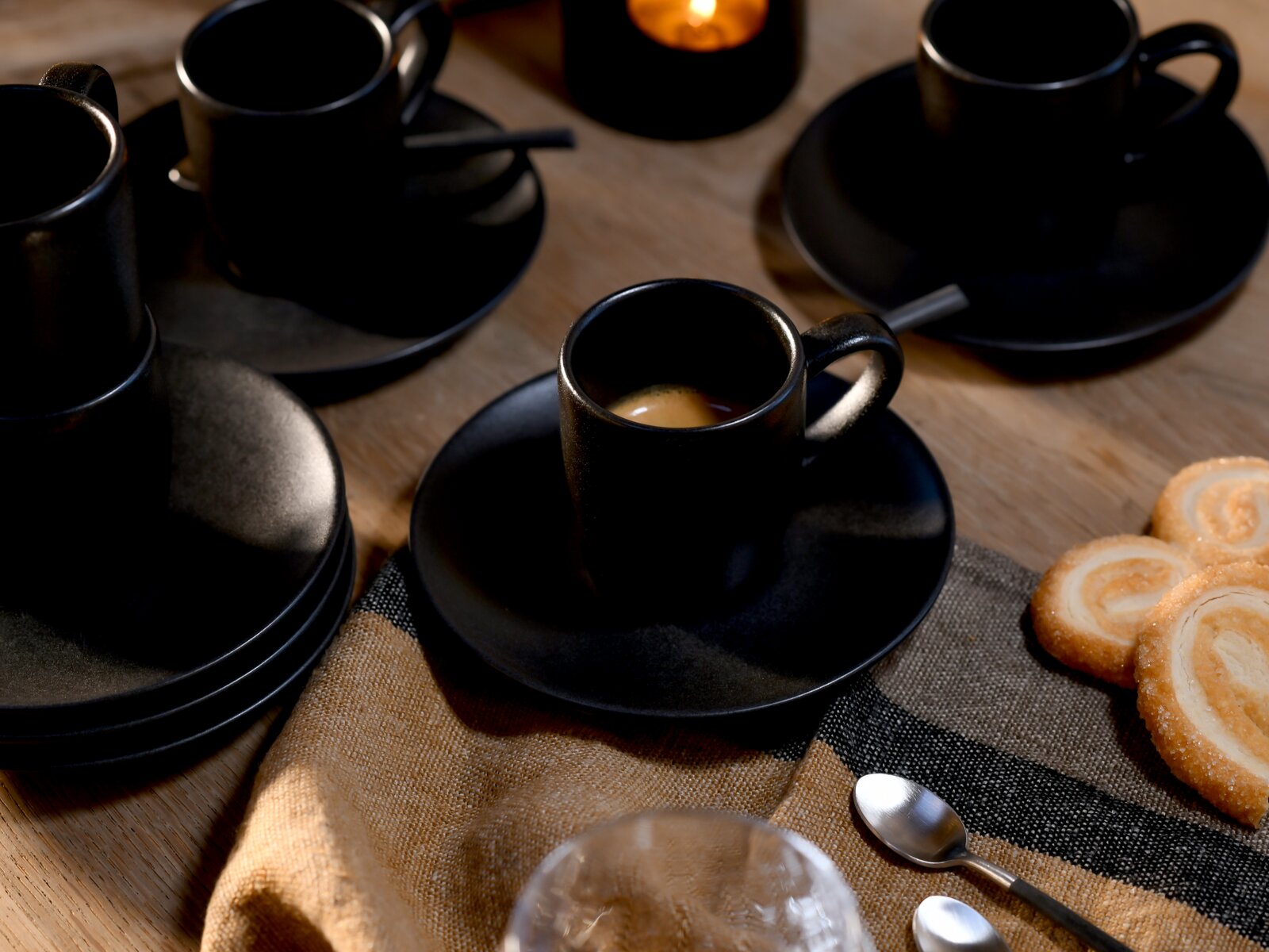 Schwarze Espressotassen mit Untertassen und Löffeln auf Holztisch, vordergründig eine Tasse mit Espresso; aufgenommen aus leicht erhöhter, schräger Perspektive