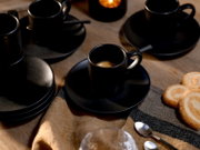 Schwarze Espressotassen mit Untertassen und Löffeln auf Holztisch, vordergründig eine Tasse mit Espresso; aufgenommen aus leicht erhöhter, schräger Perspektive