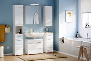 Moderne Badezimmermöbel in Weiß, bestehend aus Hochschrank, Spiegelschrank, Waschbeckenunterschrank und Hängeschrank, in einer hellblauen Umgebung mit Tageslicht von rechts.