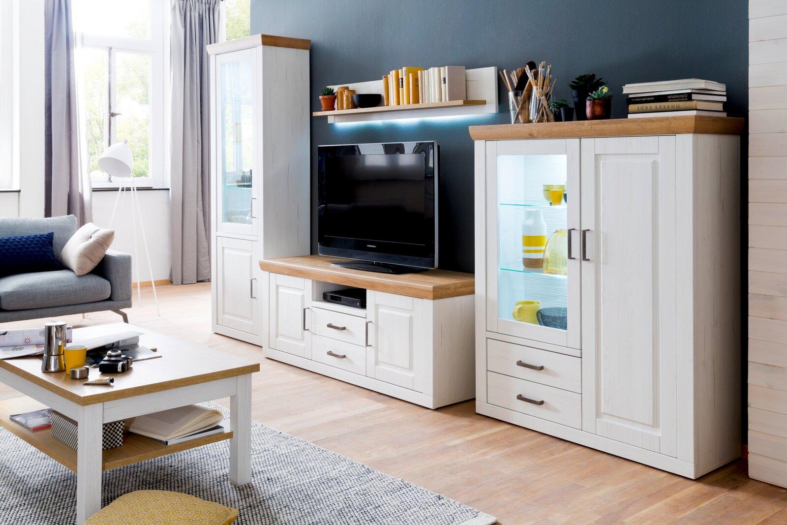 Helles Wohnzimmer mit weißer Wohnwand im Landhausstil mit Holzoberflächen, TV-Board, Vitrine mit Glasfach und Wandregal, frontal aus leicht schräger Perspektive aufgenommen