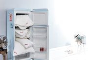 Ein geöffneter Kühlschrank mit mehreren Paradies Sommerbettdecken und Kissen, die ordentlich gestapelt sind. Die Perspektive zeigt den Kühlschrank von vorne, mit einem hellen, minimalistischen Hintergrund.