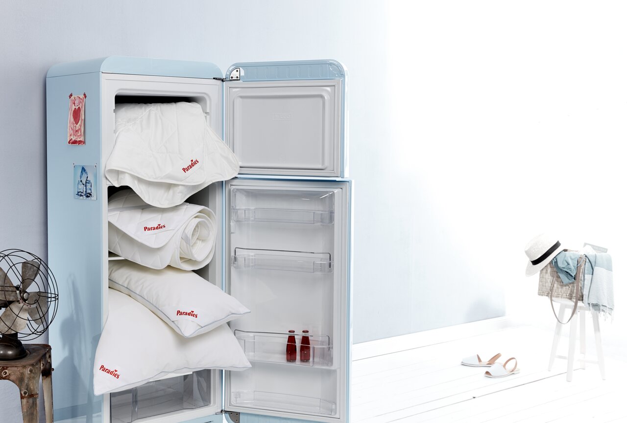 Ein geöffneter Kühlschrank mit mehreren Paradies Sommerbettdecken und Kissen, die ordentlich gestapelt sind. Die Perspektive zeigt den Kühlschrank von vorne, mit einem hellen, minimalistischen Hintergrund.