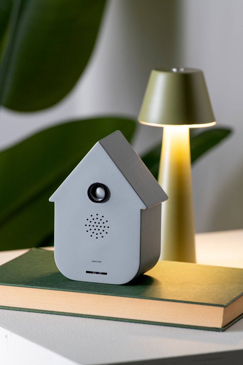 KARLSSON Geluidsbox CUCKOO Grijze geluidsbox in de vorm van een huisje, vooraan op een boek geplaatst, met een lamp op de achtergrond.
