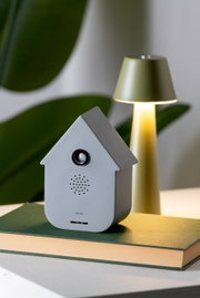 Graue Soundbox in Form eines Hauses, frontal auf einem Buch platziert, mit einer Lampe im Hintergrund.