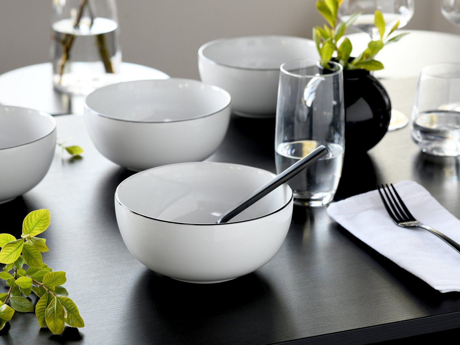 Schlichtes weißes Schüssel-Set mit feinem dunklem Rand auf gedecktem Tisch mit Glas, Besteck und schwarzer Vase, Perspektive: leicht schräg von oben (3/4-Ansicht)