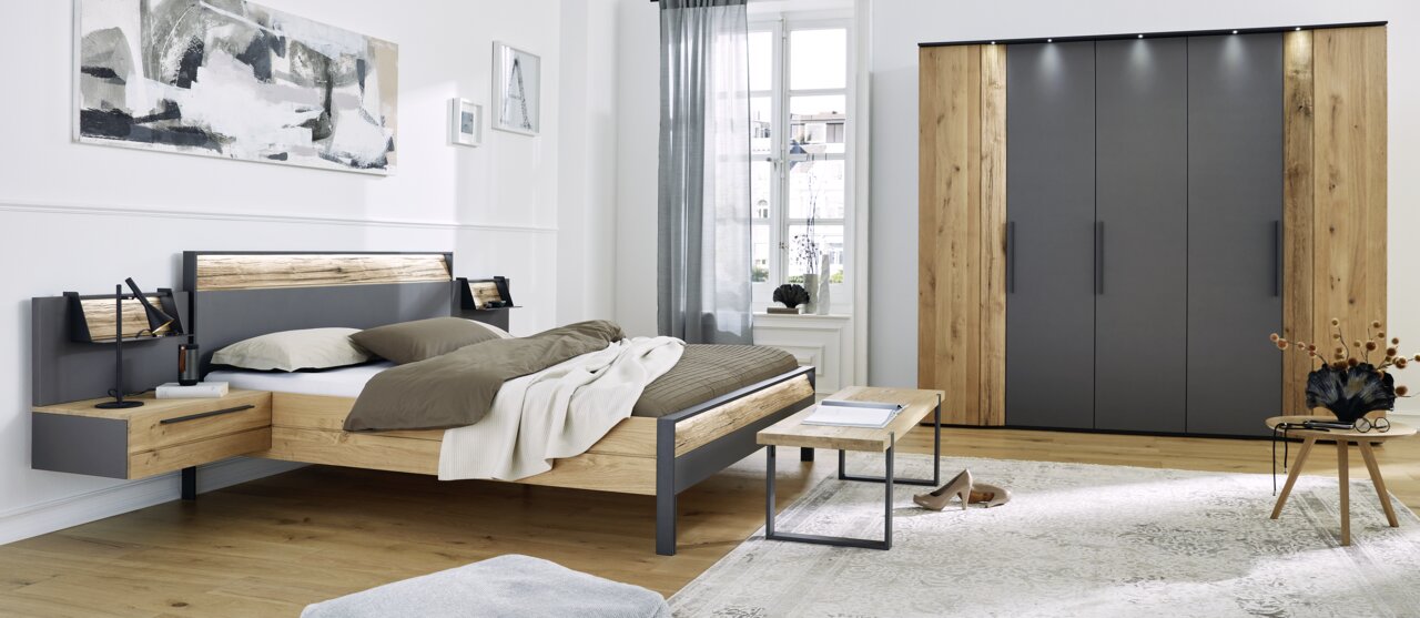 Modernes Schlafzimmer mit einem Holzbett und Nachttischen, einem großen Kleiderschrank und einem Fenster im Hintergrund. Perspektive von der Seite des Bettes.