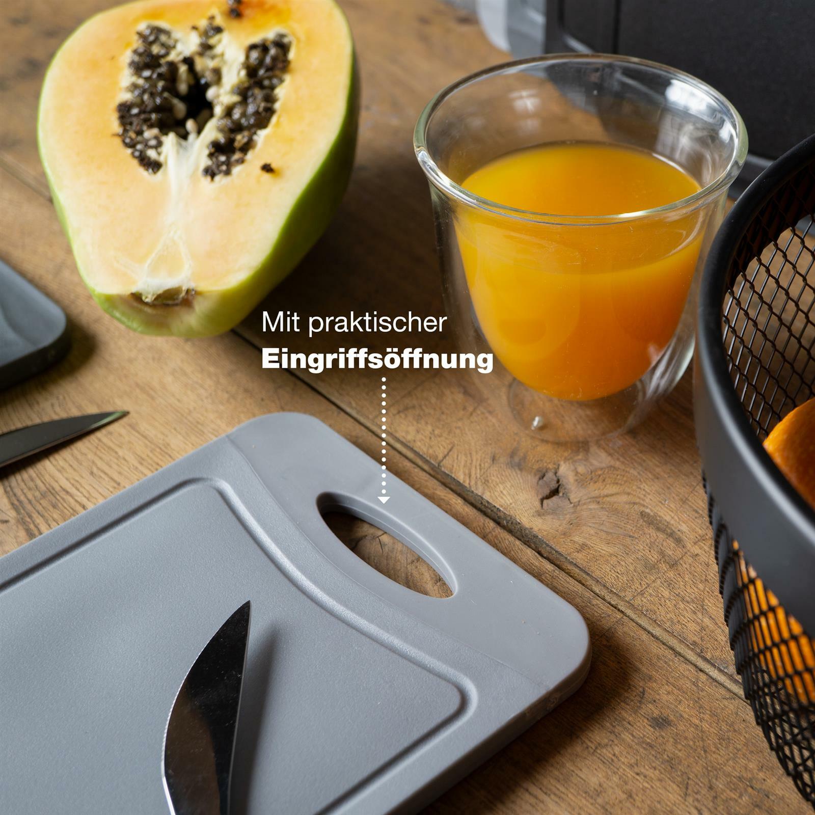 Graues Schneidebrettchen mit Eingriffsöffnung, auf einem Holztisch liegend, neben einem halbierten Papaya und einem Glas Orangensaft, aus der Vogelperspektive.