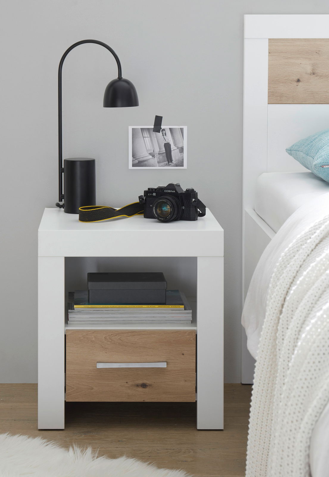 Moderne Nachtkommode in Weiß mit Holzschublade, seitliche Perspektive, dekoriert mit Lampe, Kamera und Büchern.