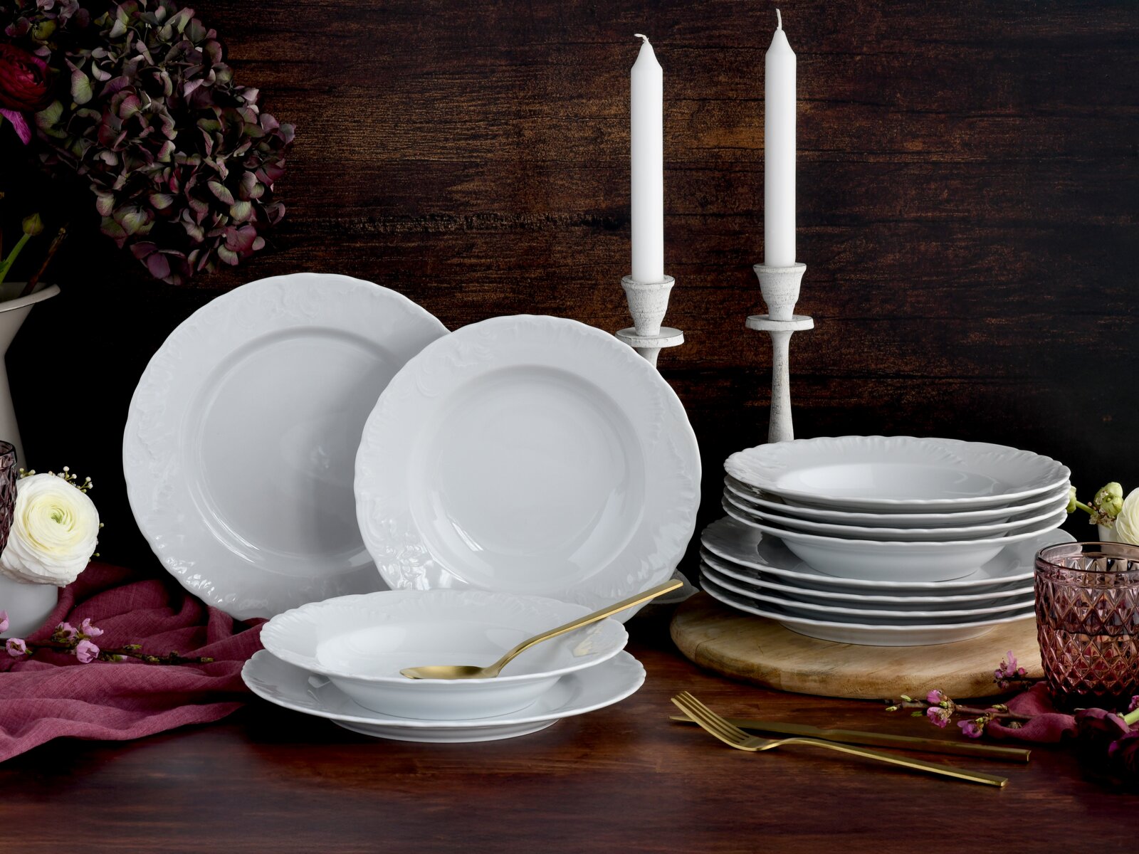 Weißes Porzellan-Geschirrset mit Reliefrand (Teller und Schalen) auf dunklem Holztisch dekoriert mit Kerzen, Blumen und goldenem Besteck, fotografiert in leicht erhöhter Frontansicht.