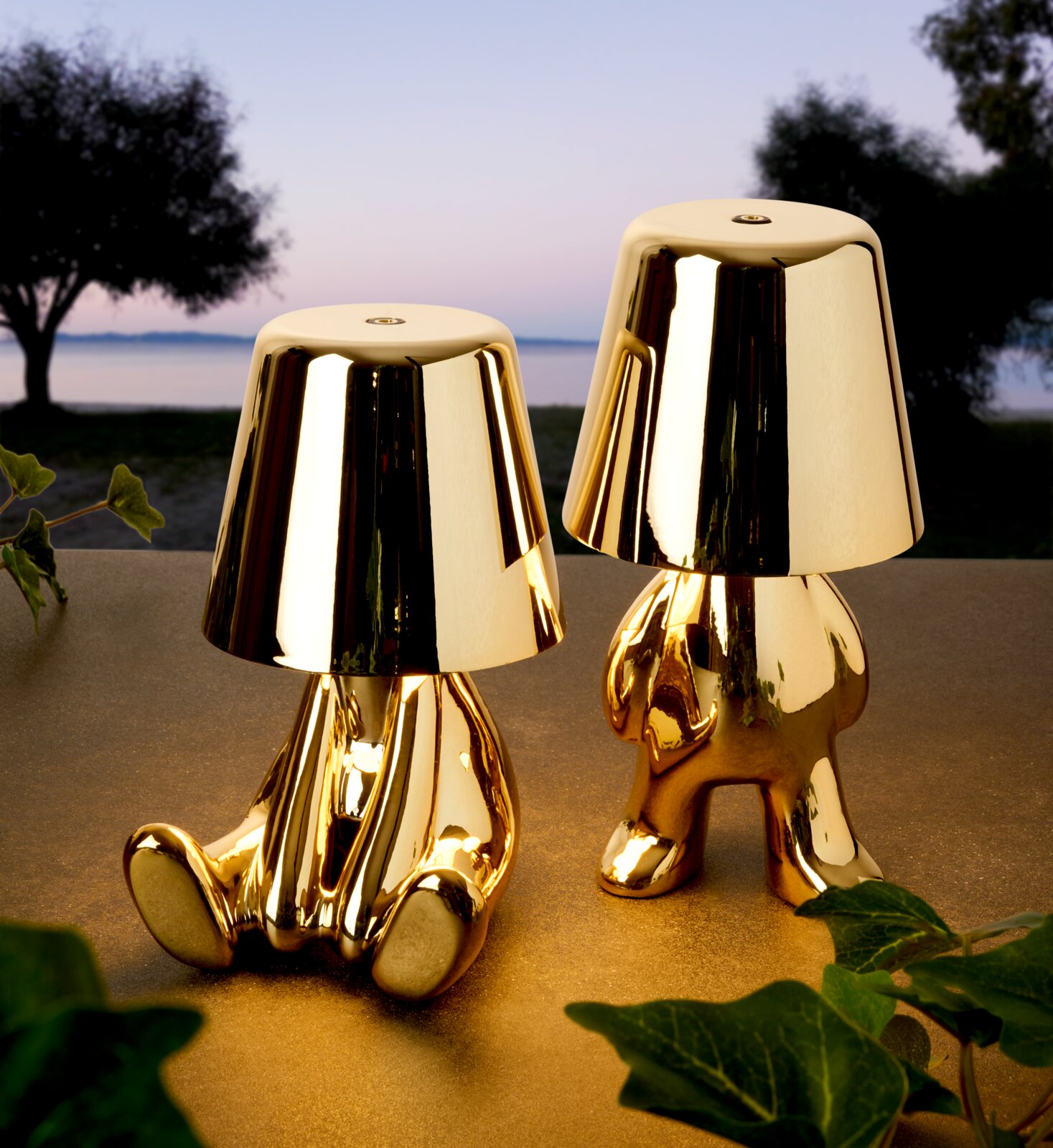 Zwei glänzende, goldfarbene Designer-Tischlampen in Figurenform auf einer Terrasse vor See- und Baumkulisse bei Dämmerung, Frontalansicht auf Augenhöhe