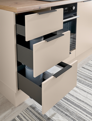 Moderne Küchenunterschränke in Beige mit Holzarbeitsplatte und drei teilweise ausgezogenen Schubladen neben einem Einbaubackofen, schräg von vorn aufgenommen