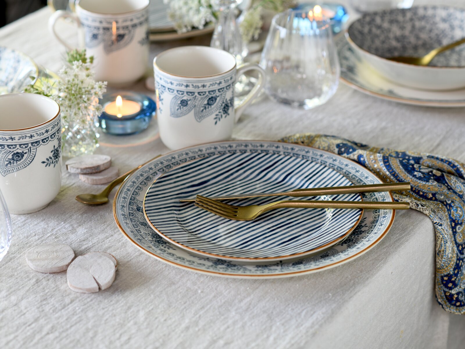 Stilvoll gedeckter Tisch mit blau-weiß gemustertem Teller-Set und Tassen, goldfarbenem Besteck und Kerzenlicht, fotografiert aus leicht schräger Vogelperspektive von oben.